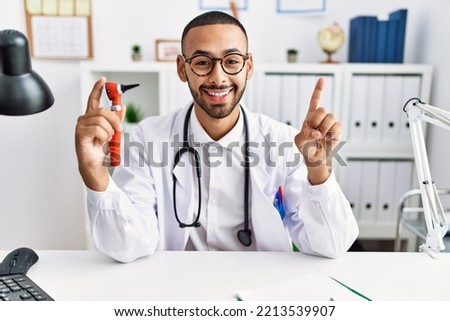 African american doctor man holding ear otoscope at the clinic smiling with an idea or question pointing finger with happy face, number one 