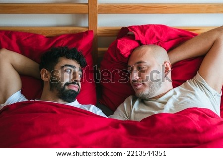 Young interracial homosexual male couple in red  bed. Mixed race and Caucasian gay  leisure time together in bedroom. Lying on back, hands under heads, facing each other with a smile. LGBT concept.  Royalty-Free Stock Photo #2213544351