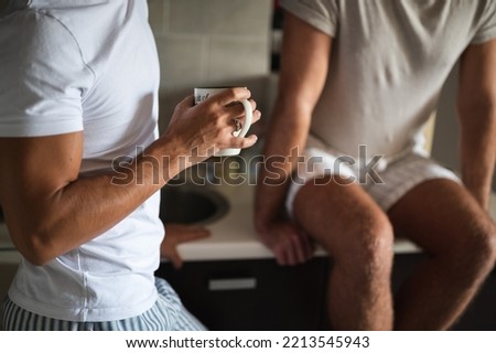 Young unrecognizable  interracial gay couple in pajamas having tea in a kitchen. Handsome homosexual men, Caucasian and Mixed race, sitting on counter, enjoy morning at home. LGBT family  concept.  Royalty-Free Stock Photo #2213545943