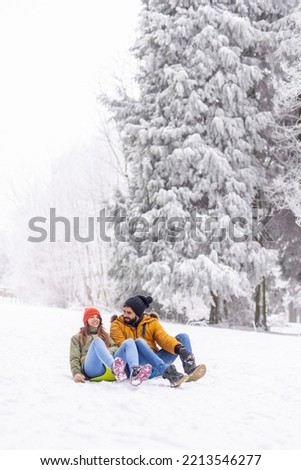 Beautiful young couple in love having fun spending winter vacation in the mountains, sliding down the hill in the snow