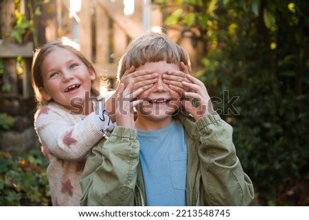 Smiling girl closes eyes to her brother. Happy kids. Make a surprise. Portrait of children in sun light. Blonk funny child play outdoor