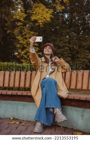 Happy woman in stylish cap and trench taking selfie on mobile phone while sitting on bench in park in fall season and having fun 