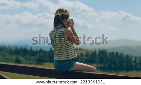 Young photographer shooting landscape on beautifull green hillway using digital camera, sitting on wooden fence, taking pictures of nature. Girl filling the portfolio with amazing photos.