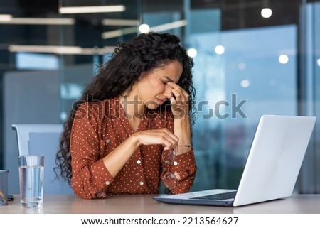 Tired business woman working inside modern office building, worker rubbing eyes with glasses off, latin american woman with curly hair using laptop for work Royalty-Free Stock Photo #2213564627