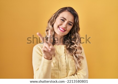 Young caucasian woman standing over yellow background smiling looking to the camera showing fingers doing victory sign. number two. 