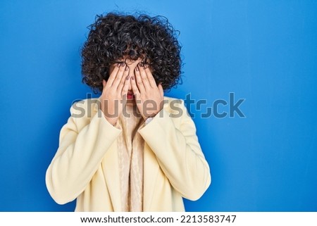 Young brunette woman with curly hair standing over blue background rubbing eyes for fatigue and headache, sleepy and tired expression. vision problem 