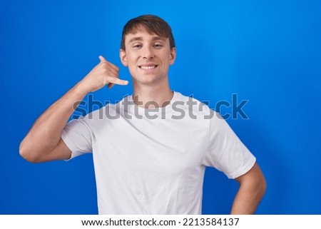 Caucasian blond man standing over blue background smiling doing phone gesture with hand and fingers like talking on the telephone. communicating concepts. 