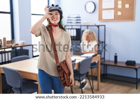 Young brunette woman working at the office wearing bike helmet smiling happy doing ok sign with hand on eye looking through fingers 