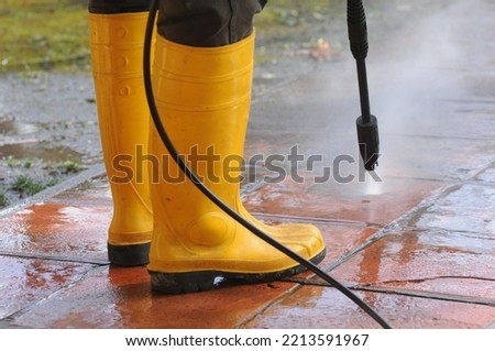 Person wearing yellow rubber boots with high-pressure water nozzle cleaning the dirt in the tiles Royalty-Free Stock Photo #2213591967
