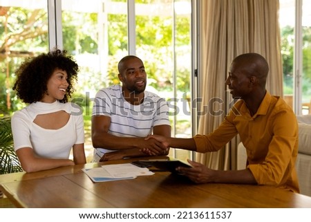 Front view of a mixed race couple sitting in their living room shaking hands with an African American male financial adviser, with sun shining in the garden outside 