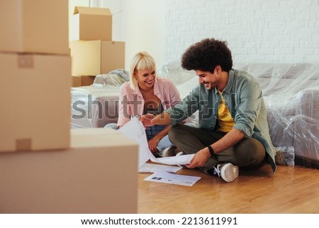 A young happy couple is moving into their new apartment and are signing the rent documents. They are sitting on the floor as they're belongings are still unpacked around them.