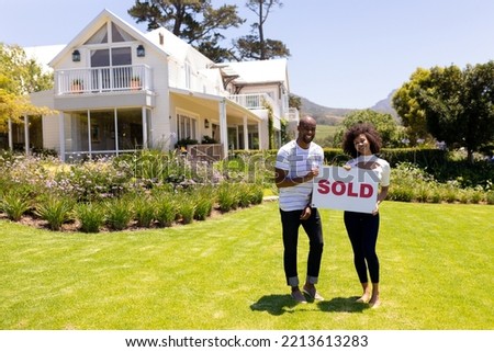 Portrait of a mixed race couple standing in the garden outside their luxury house, smiling to camera and holding a sign with the word sold written on it 