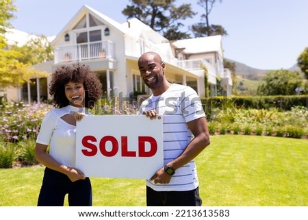 Front view of a mixed race couple standing in the garden, smiling to camera and holding sign with the word sold written on it 