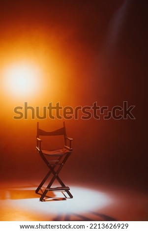 The director's chair stands in a beam of light with an orange backlight and smoke. Place for text. Free chair. Concept of selection and casting. Shadow and light. Royalty-Free Stock Photo #2213626929