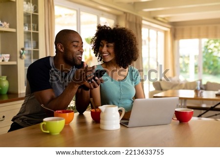 Front view of a  couple at home sitting in the kitchen, smiling at each other and using mobile phone and a laptop  Royalty-Free Stock Photo #2213633755