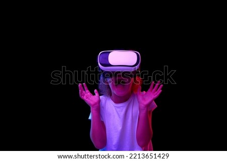 Shock. School age girl in white t-shirt enjoying 3D technology in virtual reality headset isolated on dark background in neon. Concept of cutting edge technology, video games, innovation and ad.