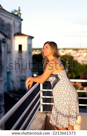 A woman on the porch of her house in light clothes looking at the street against the sky and church in the background. City of Valença, Bahia, Brazil.
