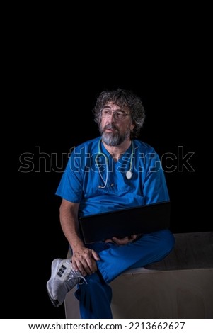 bearded mature doctor dressed in blue operating room suit sitting on a wooden box consulting a computer healthcare medical professions