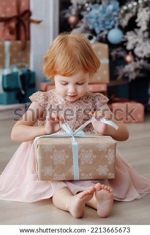 Charming little girl in a pink dress is sitting on the floor at home. She opens the Christmas present thoughtfully and looks at it.