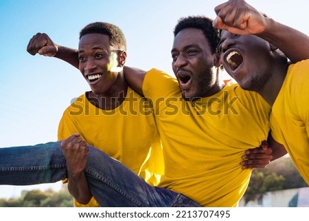 Crazy football fans celebrating in the crowd while watching the game at sport stadium - Soccer supporters screaming for their yellow team during world champion event - Focus on center African man   Royalty-Free Stock Photo #2213707495
