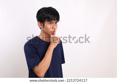 Young asian man standing while holding a finger on his lips over white background