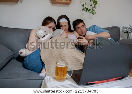 Three young friends, one boy and two girls, watching a horror movie on the laptop, screaming, scared.