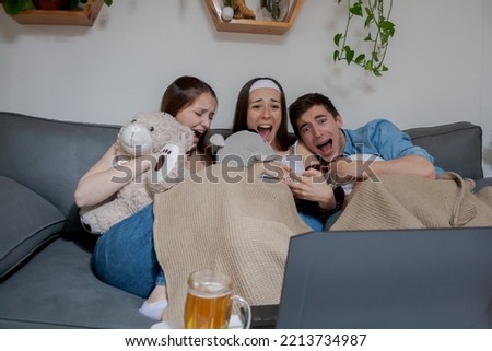 Three young friends, one boy and two girls, watching a horror movie on the laptop, screaming, scared.