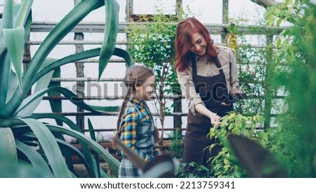 Female worker of greenhouse and her adorable little daughter are looking at plants and touching them in hothouse, woman is holding tablet. Family and gardening concept.