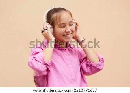 Cheerful teenager girl in a pink shirt enjoys music in headphones, isolate on a peach background