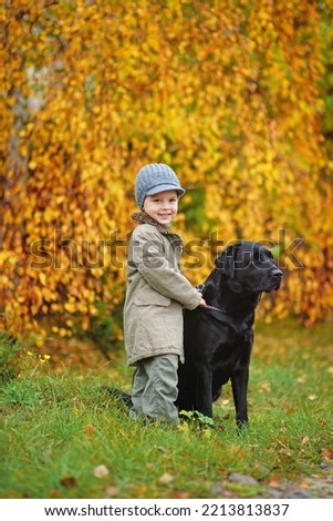               A happy little boy stands with his buddy black big labrador retriever in a beautiful autumn forest, a child plays with a dog during a walk in nature after the rain. Autumn, November     