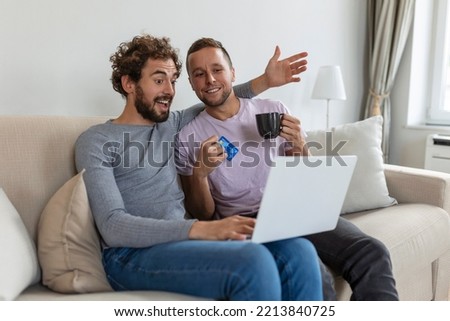 Cheerful young gay couple smiling cheerfully while shopping online at home. Two young male lovers using a credit card and a laptop to make a purchase online. Young gay couple sitting together indoors.