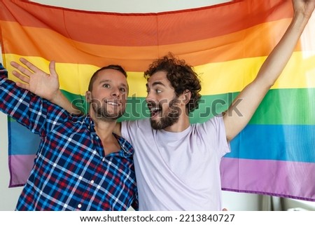 Two young gay lovers looking at each other affectionately. Two young male lovers standing together against a pride flag. Affectionate young gay couple sharing a romantic moment together.