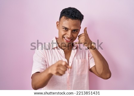 Young hispanic man standing over pink background smiling doing talking on the telephone gesture and pointing to you. call me. 