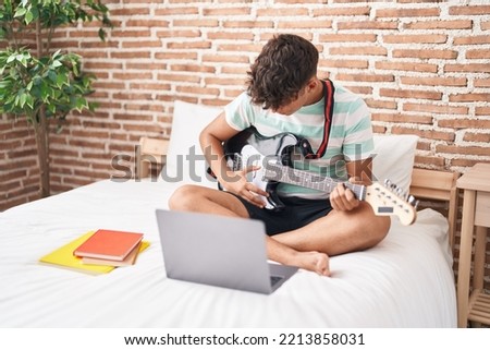 Young hispanic teenager having online electrical guitar class sitting on bed at bedroom Royalty-Free Stock Photo #2213858031