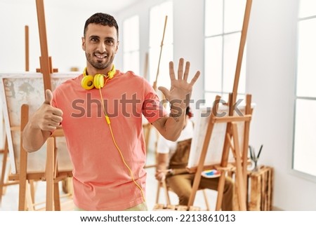 Young hispanic man at art studio showing and pointing up with fingers number six while smiling confident and happy. 