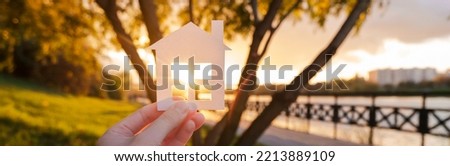 Banner with hand that holds a paper house against the background of the sunset sky. The sun is setting over the horizon. The concept of your own home, personal space and mortgage. High quality photo