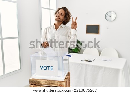 Young hispanic man voting putting envelop in ballot box smiling looking to the camera showing fingers doing victory sign. number two. 