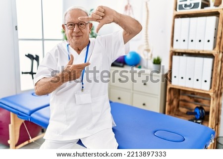 Senior physiotherapy man working at pain recovery clinic smiling making frame with hands and fingers with happy face. creativity and photography concept. 