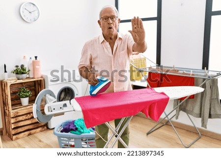 Senior man ironing clothes at home with open hand doing stop sign with serious and confident expression, defense gesture 