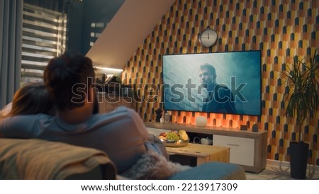 Couple on sofa in living room, watching action movie on TV or criminal blockbuster on streaming service, talking and discussing acting, resting at home on weekend. Home theater in modern apartment.