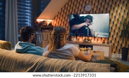 Happy couple sitting on sofa, watching action movie on TV or criminal blockbuster on streaming service, talking and discussing acting, eating snacks, pizza, chips. Spouses resting at home in evening. Royalty-Free Stock Photo #2213917331