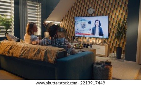 Couple sitting on sofa in living room, talking, watching TV news about poliovirus and water pollution. Man and woman spending leisure time on weekend at home together. TV live broadcasting. Royalty-Free Stock Photo #2213917449
