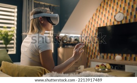 Woman wearing VR headset, sitting on sofa in cozy living room in midday, gesturing, playing video game in virtual reality, testing VR goggles, spending weekend at home. Cyberspace and metaverse. Royalty-Free Stock Photo #2213917489