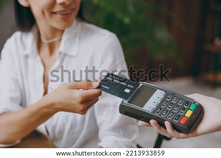 Croped young smiling fun happy woman wearing white shirt hold wireless bank payment terminal to process acquire credit card sit alone at table in coffee shop cafe restaurant indoor. Focus on machine Royalty-Free Stock Photo #2213933859