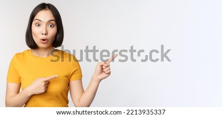 Amazed young asian woman, showing advertisement aside, pointing fingers right at promotion text, brand logo, standing happy against white background