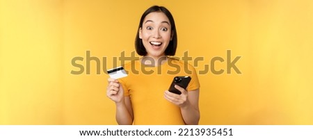 Online shopping. Cheerful asian girl holding credit card and smartphone, paying, order with mobile phone, standing over yellow background