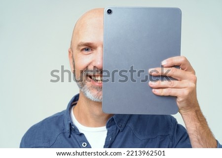 Handsome bold man cover half of his face with digital tablet in hand smiling looking at camera isolated on white background. Happy freelancer man with tablet. Using modern technologies bald man. 