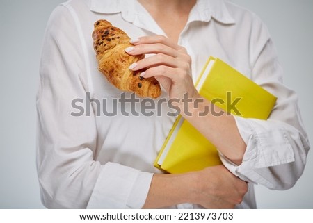Woman holding croissant and yellow book on white shirt background. Close up.