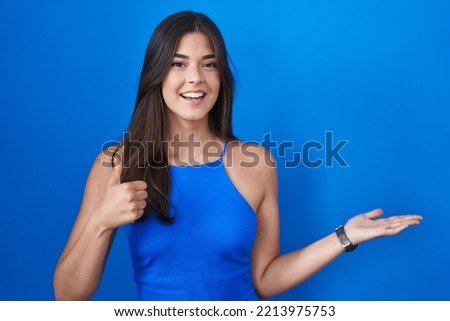 Hispanic woman standing over blue background showing palm hand and doing ok gesture with thumbs up, smiling happy and cheerful 