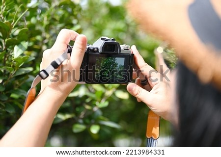 A female tourist wearing a brown straw hat uses a digital camera to photograph the beauty of the green trees in the park. Woman holding a small camera photographing leaves on blurred nature background